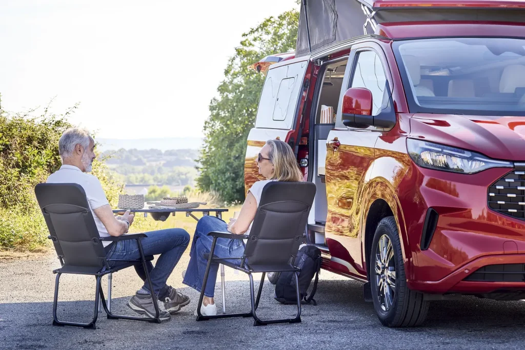 Furgoneta camper Dreamer Cap Land roja con el techo elevable abierto y una pareja sentada fuera, en la mesa y las sillas del kit de exterior